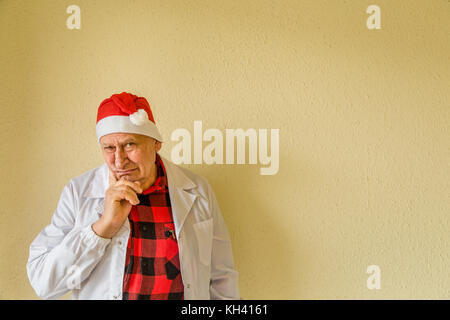 Trauriger alter Arzt mit weißem medizinischen Kleid und kariertem Hemd tragen Santa Claus hat Stockfoto