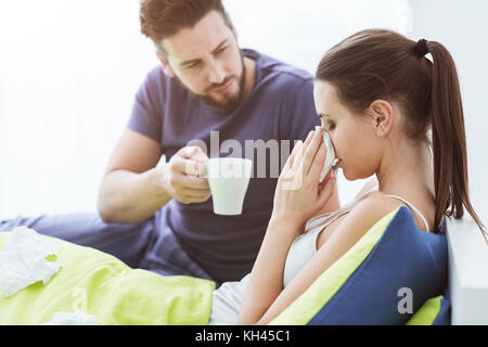 Junge Frau im Bett mit Erkältung und Grippe, sie bläst die Nase, ihrem Freund gibt ihr ein heißes Getränk und kümmert sich um Ihre Stockfoto