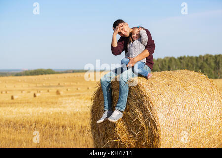 Gerne Vater und 2-jährige Tochter sitzen auf Heu Ballen in geernteten Feld Stockfoto