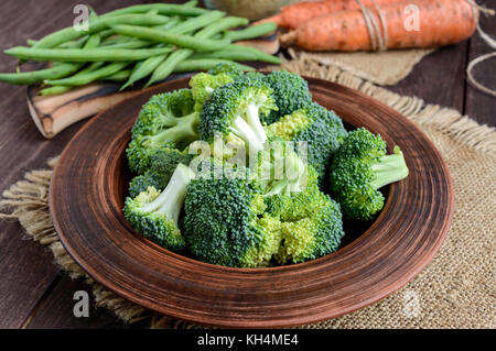 Brokkoli (Kohl) Stücke in einem Ton Schüssel auf einem hölzernen Hintergrund mit Spargel, Reis und Möhren im Hintergrund. Satz von Produkten für vegane Gerichte. Stockfoto