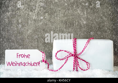 Eine weiße Geschenk mit Label mit deutschem Text frohe Weihnachten bedeutet, frohe Weihnachten. Grau grungy Zement Hintergrund mit Schnee und Schneeflocken Stockfoto