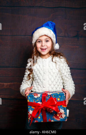 Glückliche kleine lächelnde Mädchen mit Weihnachtsgeschenk. Weihnachten Konzept. Lächelnd lustig Mädchen in Santa Hut auf Holz- Hintergrund. Stockfoto