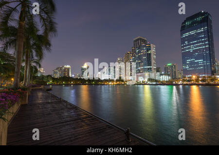 Malerischer Blick auf Palmen, Holzsteg und See an der benjakiti (benjakitti) Park und beleuchteten Wolkenkratzer in Bangkok, Thailand, am Abend. Stockfoto