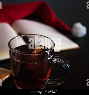 Planung zukünftiger auf Weihnachten Konzept: ein Notebook mit leeren Seiten, einen schwarzen Stift, Santa Hut, Glas Becher mit Teebeutel, alle auf dunklen Esstisch Stockfoto