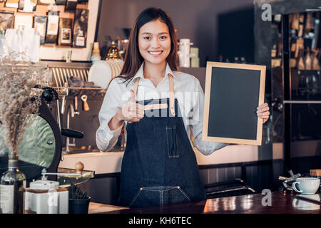 Asiatische Frau barista Schürze tragen Jean Daumen oben auf leere Tafel/Menü counter Bar mit einem Lächeln im Gesicht, cafe Service Konzept, Business Owner star Stockfoto