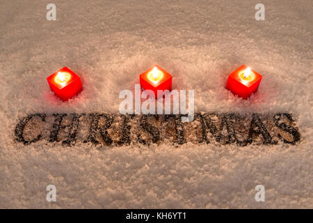 Das Wort Weihnachten in Holz- Buchstaben auf einem Schnee Hintergrund durch drei rote Kerzen beleuchtet. Stockfoto
