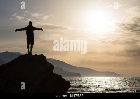 Mann mit Arme ausgestreckt feiern oder beten in schönen inspirierenden Sonnenaufgang mit Berge und Meer. Mann, Wandern oder Klettern mit Händen, genießen Sie in Stockfoto