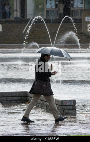 Mann in der schweren Regen, holding Dach & Phone, vorbei an den Spiegel Pool & Brunnen - Bradford City Park, West Yorkshire, England, UK. Stockfoto