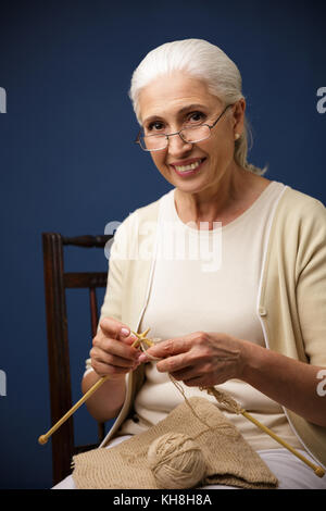 Foto der freundliche alte Frau sitzt auf dunkelblauem Hintergrund beim Stricken. Suchen Kamera. Stockfoto