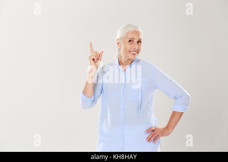 Fröhliche grauhaarige alte Dame in blauem Shirt und zeigt mit dem Finger nach oben, auf weißem Hintergrund Stockfoto