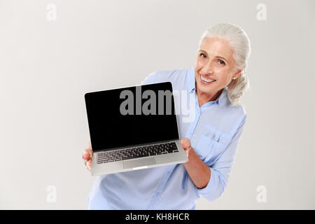 Wunderschöne reife grauhaarige Frau lächelnd und mit leerer Bildschirm des Laptop Computer isoliert Stockfoto