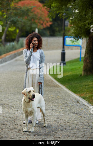 Junge besetzt schöne afrikanische Dame in casual Wandern mit Hund und am Telefon sprechen in Park Stockfoto