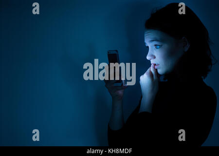 Cyber stalking: Eine junge Braunhaarige kaukasischen teenage Mädchen auf ihrem Mobiltelefon, Smartphone, Handy, besorgt suchen, verwirrt, besorgt durch den Inhalt der Botschaft, die sie ist zu sehen, in der Nacht, Großbritannien Stockfoto