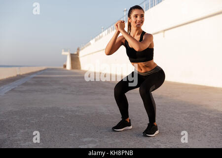 Junge schöne kaukasischen Dame in Sportswear und sitzt - Übung während der Ausbildung in der Nähe von Meer in Morgen Stockfoto