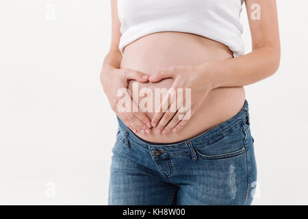 Zugeschnittenes Bild der schwangeren Frau, die ihre Hand auf den Bauch im Herzen Geste auf weißem Hintergrund Stockfoto