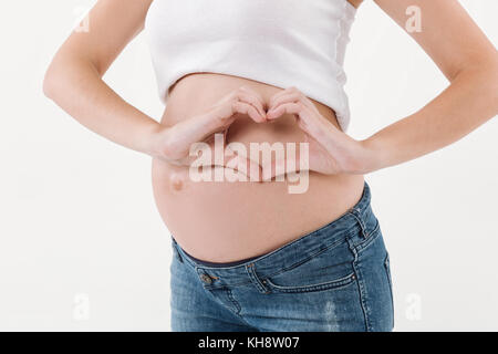 Zugeschnittenes Bild der schwangeren Frau, die ihre Hand auf den Bauch und Herz Geste auf weißem Hintergrund Stockfoto