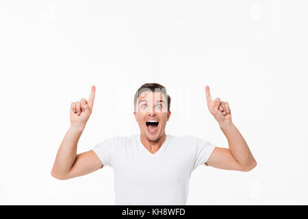 Portrait einer amüsiert aufgeregt Mann in einem weißen T-Shirt stand und zeigen Sie mit zwei Fingern auf Platz kopieren, während Sie die Kamera über Weiß ba isoliert Stockfoto