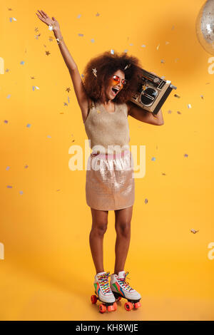 In voller Länge Porträt der Schönen überglücklich afro-amerikanische disco Frau mit erhobenen Hand, stehend auf Rollschuhen, Boombox, isoliert auf Gelb Stockfoto