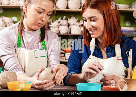 Weibliche potter lehrt Frau in einem jeanshemd und Schürze macht ein Loch zu machen, eine tiefe Schüssel auf dem Hintergrund Regal mit Lehm Produkte Stockfoto