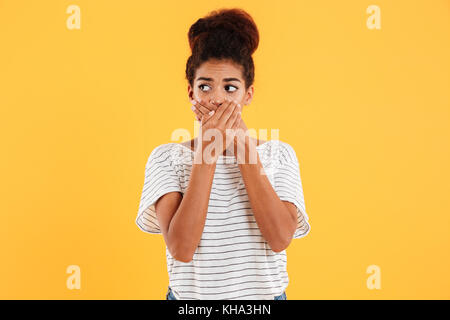 Junge nervöse afrikanische Frau, die ihren Mund und beiseite sucht über gelb isoliert Stockfoto
