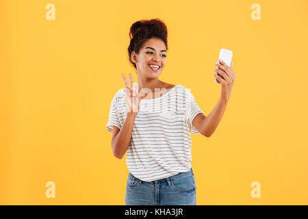 Afrikanische positive hübsche Dame, selfie auf Smartphone und zeigt Frieden Geste isoliert Stockfoto