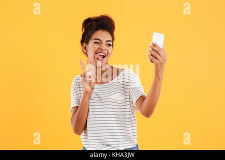 Junge lustig afrikanischen Casual lady Grimassen und selfie auf dem Smartphone über gelb isoliert Stockfoto