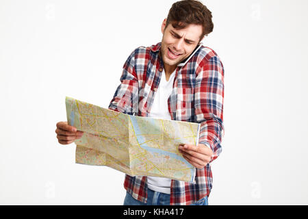 Porträt eines verwirrten jungen Mann Gespräch am Handy, während im Travel map Über weißem Hintergrund suchen Stockfoto
