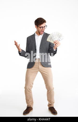 Volle Länge Portrait von fröhlichen aufgeregt Mann in Brillen und eine Jacke holding Bündel geld Banknoten im Stehen und feiert über isoliert Stockfoto