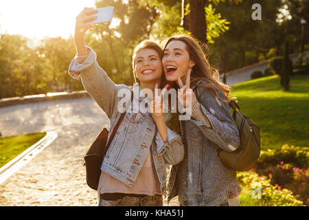 Nahaufnahme von zwei hübsche junge Frau in Freizeitkleidung, Frieden Geste beim Bilden von selfie auf dem Smartphone, outdoor Stockfoto