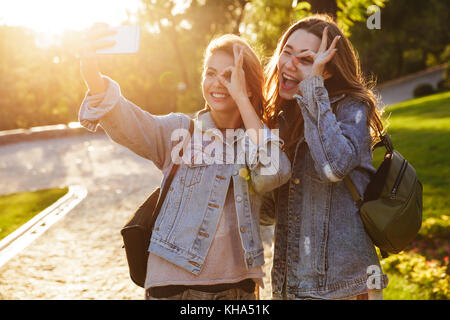 Nahaufnahme von zwei lustige junge Brünette in Freizeitkleidung durch OK Zeichen beim Bilden von selfie auf dem Smartphone, outdoor Stockfoto