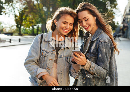 Zwei glückliche junge brünette Frau in Jeans Jacke Hören von Musik auf Handy beim Gehen auf die Straße Stockfoto