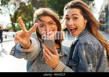 Zwei glückliche jugendlich Mädchen zeigen Frieden Zeichen beim Hören von Musik auf dem Smartphone, Kamera suchen, Außenpool Stockfoto