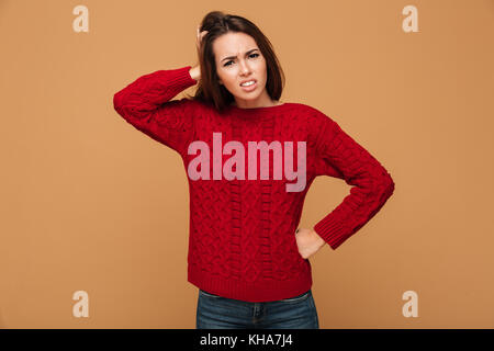 Frustrierte junge brünette Frau in Rot in Form gestrickter Pullover mit Hand auf ihre Hüfte, Kopf, auf beige Hintergrund isoliert Stockfoto