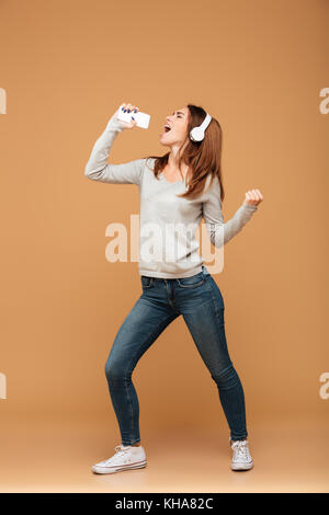 Volle Länge Foto, fröhliche junge Frau in Freizeitkleidung halten Sie Ihr Smartphone wie Mikrofon, auf beige Hintergrund isoliert Stockfoto