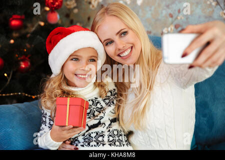 Süß lächelnde Mädchen in Santa's Hut holding Geschenkbox, während ihre Mutter unter selfie auf dem Smartphone Stockfoto