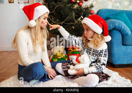 Süße kleine blonde Mädchen in Santa's Hut berühren Mütter Nase, holding Teddybär beim Sitzen auf dem weißen Teppich in der Nähe von Weihnachten Baum Stockfoto