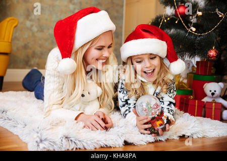 Glückliche junge Frau und ein kleines Mädchen in Santa's Hut spielen mit Snow Ball während auf weißen Teppich im Wohnzimmer liegen Stockfoto