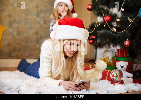 Junge hübsche blonde Frau in Santa's Hut Kontrolle Nachrichten auf dem Smartphone, während auf dem Boden liegend in der Nähe von Weihnachten Baum Stockfoto