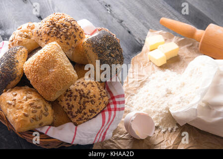 Backen Thema Bild mit einem Bündel gemischte Brötchen, in einem Weidenkorb, mit Mehl, Eiern und Butter in der Nähe von Es, auf einem schwarzen Holz- Hintergrund. Stockfoto