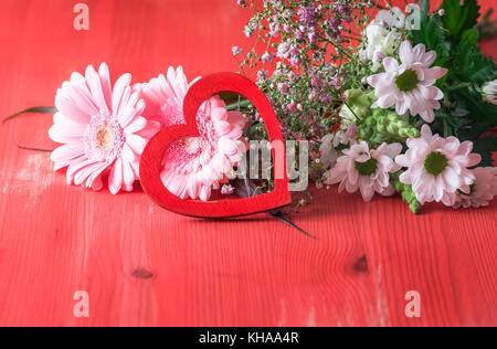 Romantische Bild mit einem Bouquet von verschiedenen Chrysantheme Blumen und eine dekorative Holz- Herzen gegen sie lehnte, dargestellt auf einem roten Holz- backgrou Stockfoto