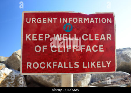 Schild am Strand Warnung über rock-Falls, wo grosse Felsbrocken gegen roten Sandsteinfelsen angehäuft wurden weitere Erosion durch das Meer zu verhindern. Stockfoto