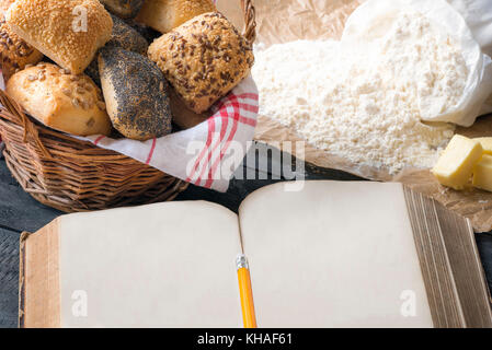 Antike offenes Buch mit Platz für Text, von frischen, hausgemachten Brötchen und Backzutaten umgeben. ein Konzept für das Backen, Kochen, Rezept. Stockfoto