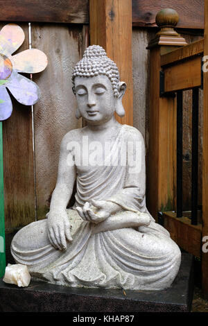 Serene Buddha Stein Statue in sitzender Position meditieren Pose auf hölzernen Veranda mit Blume im Hintergrund und Crystal im Vordergrund Stockfoto
