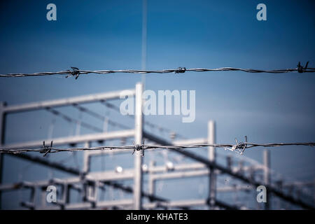 Hinter einem stacheldraht Sicherheitszaun mit Schwerpunkt auf den Zaun, mit einem Kraftwerk jenseits von Fokus mit blauem Himmel. Stockfoto