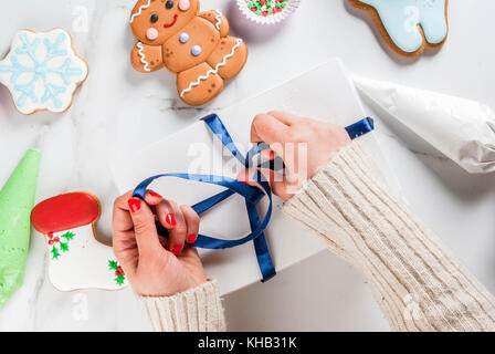 Die Vorbereitung auf Weihnachten, Verzierung traditionellen Lebkuchen mit bunten Zucker Glasur, das Mädchen faltet die Cookies in einem weißen Geschenkbox, mit Band Stockfoto