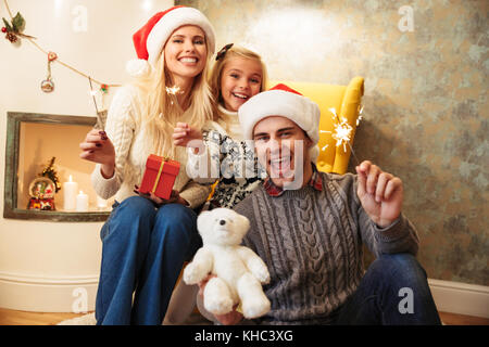 Überglücklich Familie von drei Person im Stuhl sitzen, holding Sparkler, Kamera, während wir Weihnachten feiern Stockfoto