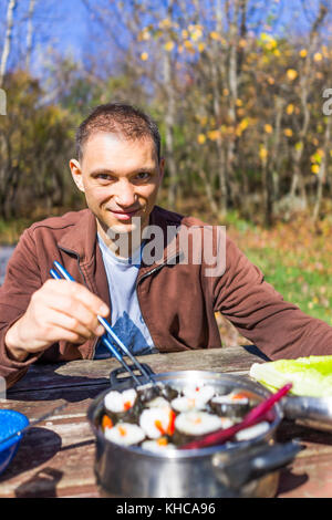 Portrait der junge Mann lächelnd sitzen auf hölzernen Picknicktisch in Campground mit Stäbchen bereit Sushi im Herbst camping Reise zu essen Stockfoto