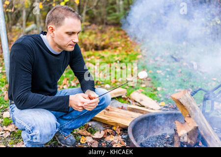 Nahaufnahme des jungen Mannes rösten eine weiße Marshmallow caramelizing auf Feuer, Detail und Textur von Campingplatz Lagerfeuer grillen im freien Park mit Stockfoto