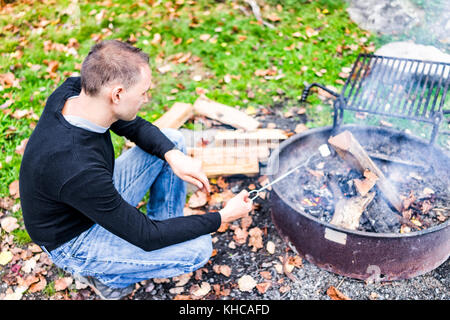 Nahaufnahme des jungen Mannes rösten eine weiße Marshmallow caramelizing auf Feuer, Detail und Textur von Campingplatz Lagerfeuer grillen im freien Park mit Stockfoto