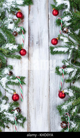 Snowy christmas tree branches, Zuckerstangen und roten Verzierungen bilden vertikale Grenzen auf rustikalen weißem Holz Stockfoto